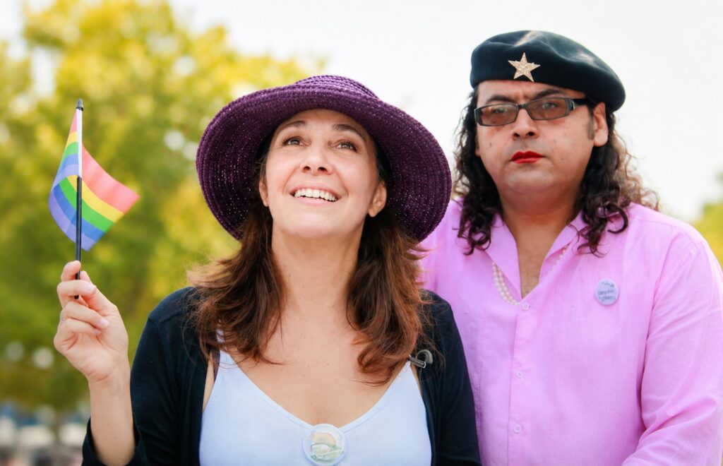 Two people at a public demonstration holding a rainbow flag, with political and identity symbols visible.