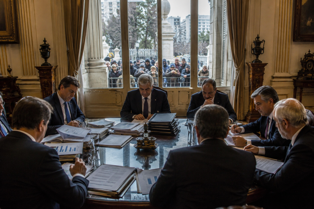 Government officials reviewing public spending documents while citizens wait outside.