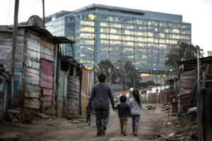 Mother and children walking through a low-income neighborhood with a large government office complex in the background, illustrating the contrast between public spending and child poverty.
