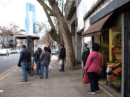 Anonymous workers at a Montevideo bus stop carrying groceries, with a modern office tower in the distance.