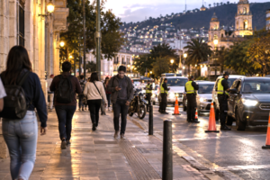 Pedestrians crossing a Latin American city street near a discreet police security checkpoint at dusk.