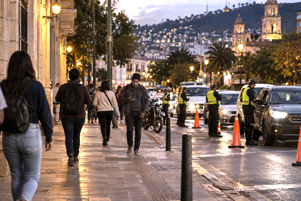 Pedestrians crossing a Latin American city street near a discreet police security checkpoint at dusk.