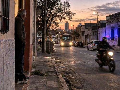 Anonymous elderly man standing at a doorway in a Montevideo neighborhood at dusk as a bus and motorcycle pass nearby.