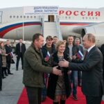 Russian officials greet returning passengers on a red carpet beside a government aircraft at an airport.
