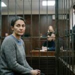 A woman seated inside a secured courtroom enclosure under the supervision of a uniformed guard in Russia.