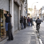 Small business owner opening a shop in Montevideo as workers pass and a public assistance queue forms nearby.