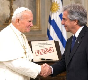 A Catholic pope shakes hands with a Uruguayan president beside a folder marked VETADO, with Uruguay’s flag in the background.